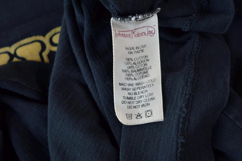 May include: A white care tag with black text that reads "Johnson Motors, Inc." and includes care instructions for the garment. The tag is attached to a dark blue t-shirt.