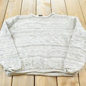 Könnte beinhalten: Ein hellgrauer Strickpullover mit Rundhalsausschnitt. Der Pullover hat ein strukturiertes Muster und lange Ärmel. Das Kleidungsstück liegt gefaltet auf einer Holzoberfläche.
