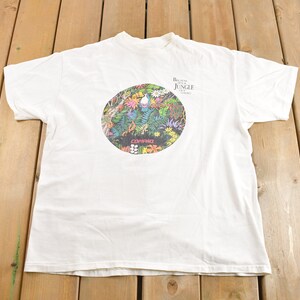May include: White t-shirt with a circular graphic of a jungle scene. The graphic features a variety of plants, flowers, and animals. The text "The Jungle Book" is printed above the graphic.