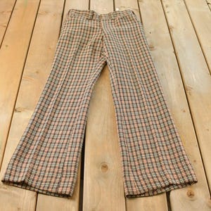May include: Brown, orange, and white plaid pants with a flared leg. The vintage trousers are laid flat on a wooden surface. The pants have a button and zipper closure.