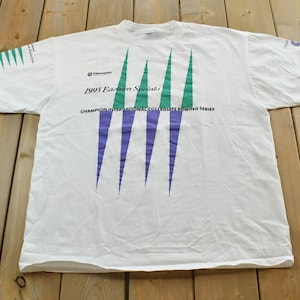 May include: White t-shirt with a Champion logo and text that reads "1995 Eastern Sprints". The shirt features a design of green and purple vertical lines. The text "Champion International Collegiate Rowing Series" is also printed on the shirt.