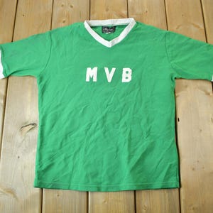 May include: Green short-sleeved t-shirt with white trim around the neck and sleeves. The shirt has the letters "MVB" in white on the front.
