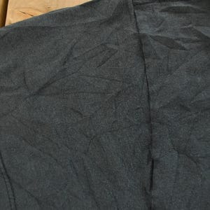 Op de afbeelding: Close-up van een zwart t-shirt, met de schouder en mouw. De stof is getextureerd en licht gekreukt. De stiksels zijn zichtbaar langs de naden. Het shirt lijkt gemaakt van een zacht materiaal.