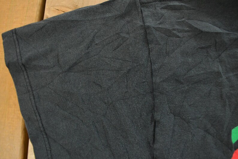 Op de afbeelding: Close-up van een zwart t-shirt, met de schouder en mouw. De stof is getextureerd en licht gekreukt. De stiksels zijn zichtbaar langs de naden. Het shirt lijkt gemaakt van een zacht materiaal.