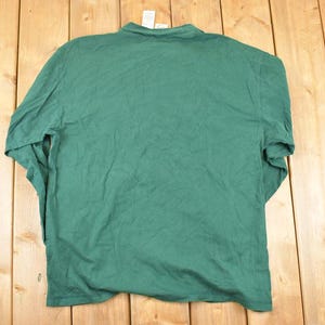 May include: A solid green long-sleeved shirt, viewed from the back, hangs against a wooden background. The shirt has a crew neck and appears to be made of a soft, lightweight fabric. The garment is slightly wrinkled, suggesting it has been worn or stored.
