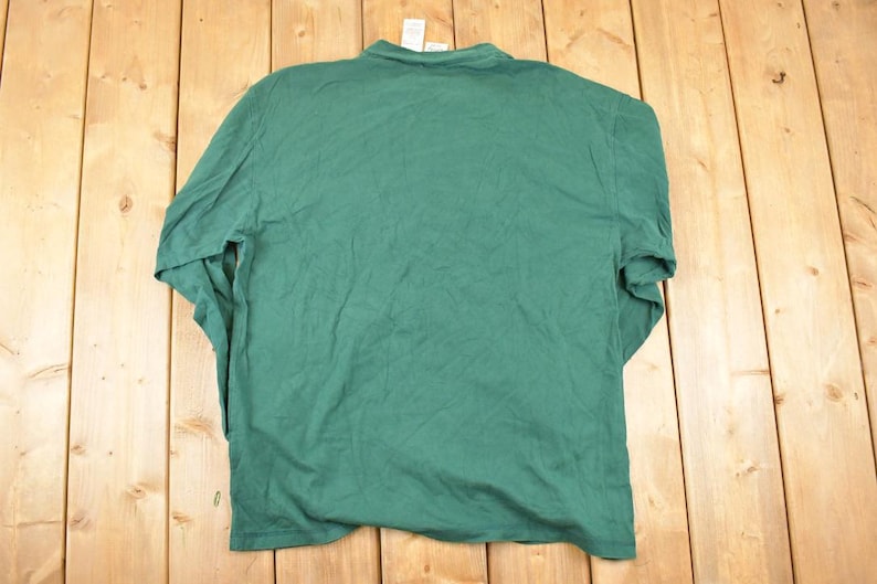 May include: A solid green long-sleeved shirt, viewed from the back, hangs against a wooden background. The shirt has a crew neck and appears to be made of a soft, lightweight fabric. The garment is slightly wrinkled, suggesting it has been worn or stored.