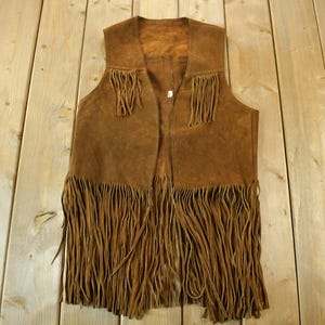May include: A brown suede vest with long fringe detailing. The vest has a classic design with fringe along the bottom and chest. The vest is a warm brown colour and appears to be vintage.