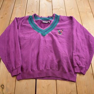 Könnte beinhalten: Lilafarbener Pullover mit einem grün-weiß gestreiften V-Ausschnitt und einem kleinen gestickten Logo auf der Vorderseite.