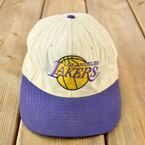 May include: A white and purple baseball cap with the Los Angeles Lakers logo embroidered on the front. The logo features a basketball with the team name and city written around it.