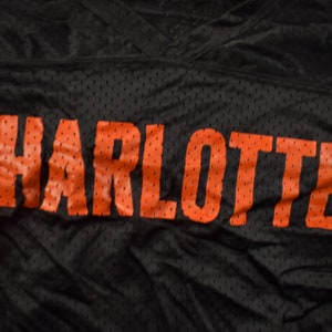 Op de afbeelding: Zwart shirt met het woord "CHARLOTTE" in oranje gedrukt.