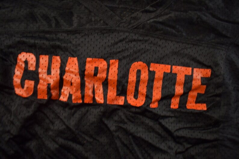 Op de afbeelding: Zwart shirt met het woord "CHARLOTTE" in oranje gedrukt.