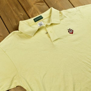 K&ouml;nnte beinhalten: Ein hellgelbes Poloshirt mit einem gr&uuml;nen Cutter & Buck-Logo am Kragen und einem gestickten roten, wei&szlig;en und schwarzen Cartoon-Tiger auf der rechten Brust.