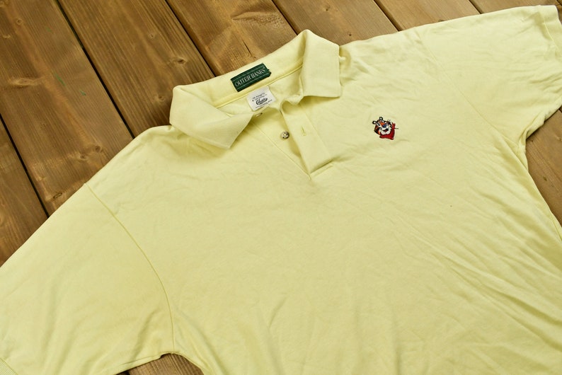 K&ouml;nnte beinhalten: Ein hellgelbes Poloshirt mit einem gr&uuml;nen Cutter & Buck-Logo am Kragen und einem gestickten roten, wei&szlig;en und schwarzen Cartoon-Tiger auf der rechten Brust.