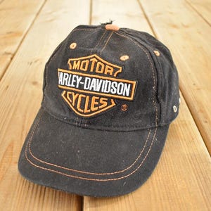 Może przedstawiać: Czarna czapka bejsbolowa z logo Harley-Davidson Cycles w kolorze pomarańczowym i białym. Czapka ma zakrzywiony daszek i pomarańczowe szwy. Wykonana z wytrzymałej tkaniny.