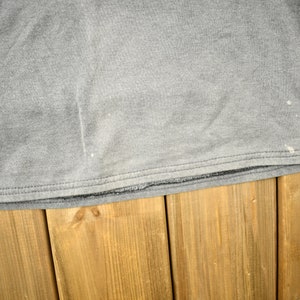 Puede incluir: Una camiseta gris de manga larga con cuello redondo. La camiseta tiene un aspecto desgastado con salpicaduras de pintura blanca. El pu&ntilde;o de la manga tiene un estampado de rayas. La camiseta est&aacute; sobre una superficie de madera.