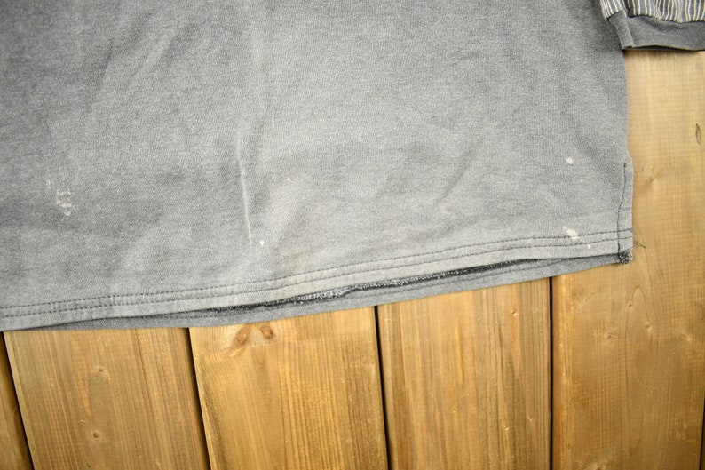 Puede incluir: Una camiseta gris de manga larga con cuello redondo. La camiseta tiene un aspecto desgastado con salpicaduras de pintura blanca. El pu&ntilde;o de la manga tiene un estampado de rayas. La camiseta est&aacute; sobre una superficie de madera.