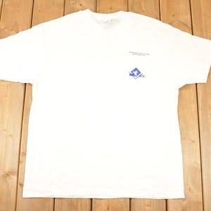 Op de afbeelding: Wit T-shirt met een ronde hals. Het shirt heeft een klein blauw logo op de linkerbovenborst. Het logo is een gestileerd ontwerp in een blauw vierkant. Het shirt ligt plat op een houten oppervlak.