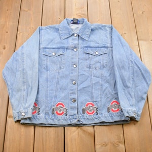 May include: A light blue denim jacket with four Ohio State logos on the bottom hem. The logos are red and gray.