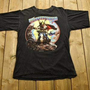 Può includere: T-shirt nera con il nome della band "MOLLY HATCHET" sopra una grafica colorata di una figura di guerriero. Il guerriero è in una scena di battaglia con una spada e un mantello. La maglietta è stesa.