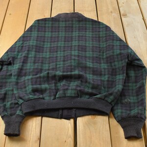 Puede incluir: Chaqueta bomber vintage con un cl&aacute;sico estampado de cuadros verdes y azul marino. La chaqueta tiene cuello, pu&ntilde;os y dobladillo de canal&eacute; negro. La chaqueta est&aacute; hecha de un material suave, posiblemente lana.