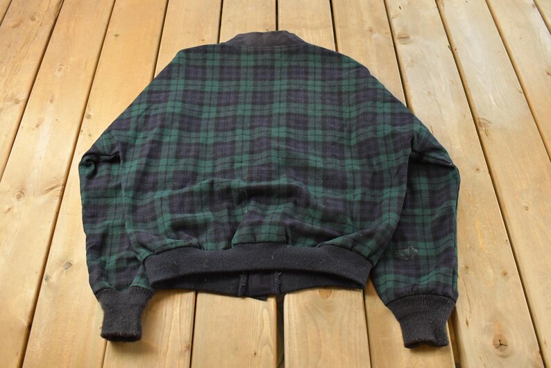 Puede incluir: Chaqueta bomber vintage con un cl&aacute;sico estampado de cuadros verdes y azul marino. La chaqueta tiene cuello, pu&ntilde;os y dobladillo de canal&eacute; negro. La chaqueta est&aacute; hecha de un material suave, posiblemente lana.