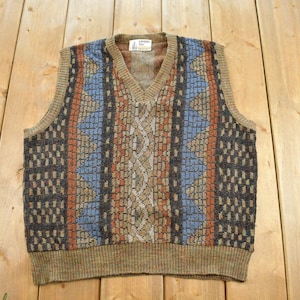 May include: A sleeveless V-neck sweater vest featuring a multi-colored geometric and textured knit pattern in shades of brown, blue, and tan. The garment has a ribbed hem and neckline, with a central vertical cable knit design and a label at the neck.