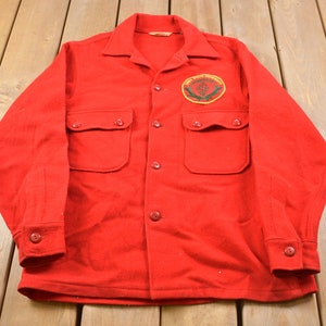 May include: Red wool shirt with a patch on the left chest that says "Conservation Department" with a green pine tree and a deer inside a circle. The shirt has two large pockets on the front and button closures.