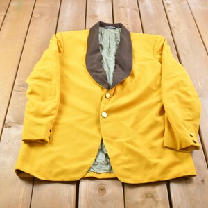 Könnte beinhalten: Ein gelber Blazer mit einem braunen Samtkragen. Der Blazer hat einen einzigen goldenen Knopf und zwei goldene Knöpfe an jedem Ärmel.