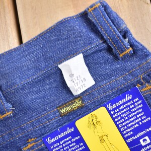 Op de afbeelding: Een close-up van een paar blauwe denim jeans met een bruine leren tailleband. De jeans heeft een label aan de tailleband dat "Wrangler" zegt en de maat informatie "9/2 17/18 4972" heeft. Er is een blauw en wit label aan de jeans bevestigd dat "Garantie" in het Frans en "Guarantee" in het Engels zegt.