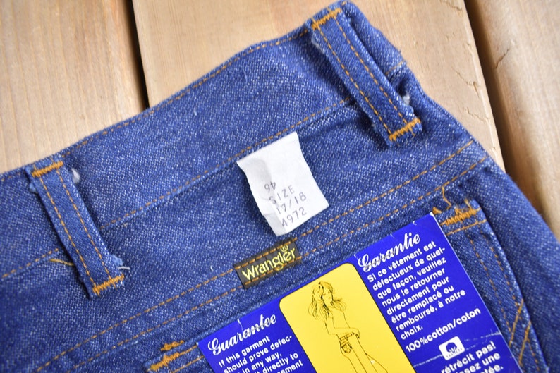 Op de afbeelding: Een close-up van een paar blauwe denim jeans met een bruine leren tailleband. De jeans heeft een label aan de tailleband dat "Wrangler" zegt en de maat informatie "9/2 17/18 4972" heeft. Er is een blauw en wit label aan de jeans bevestigd dat "Garantie" in het Frans en "Guarantee" in het Engels zegt.