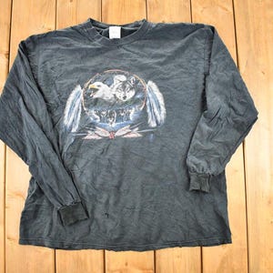 May include: Dark grey long-sleeved shirt with a graphic of an eagle, wolves, and feathers in a circular design. The shirt has a crew neck and ribbed cuffs. The graphic is in shades of white, grey, and blue.