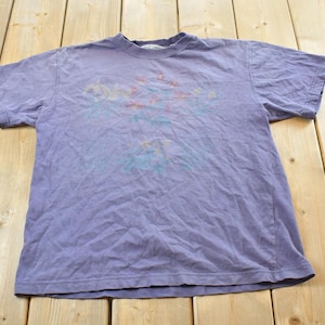 Peut inclure: Un t-shirt violet délavé à col rond et manches courtes. Le t-shirt présente un motif graphique délavé dans les tons de rouge, bleu et jaune sur le devant. Le t-shirt est posé à plat sur une surface en bois.