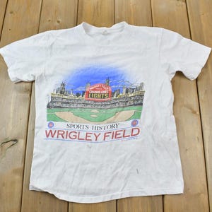 Pode incluir: Camiseta branca com um gráfico de Wrigley Field, casa dos Chicago Cubs, com o texto "Sports History Wrigley Field".