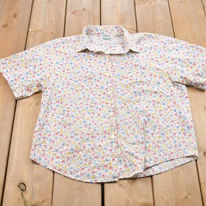 Puede incluir: Camisa blanca de manga corta con un estampado floral colorido. La camisa tiene un cuello clásico y un bolsillo en el pecho. El diseño floral incluye flores rojas, azules y amarillas.