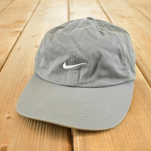 Op de afbeelding: Een grijze baseballcap met een gebogen klep en een wit Nike swoosh-logo. De pet is gemaakt van een zachte stof en ligt op een houten oppervlak.