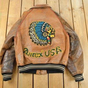May include: Brown leather jacket with a Native American chief head patch on the back. The jacket has brown sleeves and a black and white striped hem. The text "Avirex USA" is embroidered below the patch.