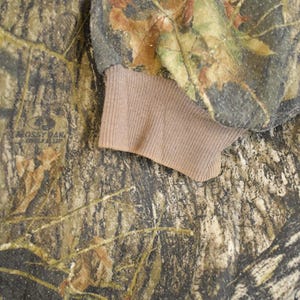 May include: Close-up of a camouflage pattern fabric with brown and green hues, featuring a ribbed tan cuff. The fabric has a detailed pattern of leaves and branches, with the "Mossy Oak" logo visible.