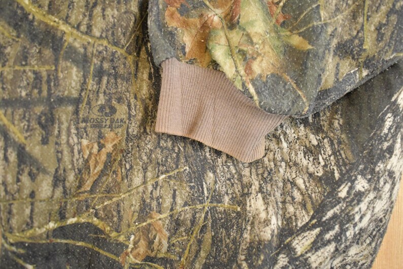 May include: Close-up of a camouflage pattern fabric with brown and green hues, featuring a ribbed tan cuff. The fabric has a detailed pattern of leaves and branches, with the "Mossy Oak" logo visible.