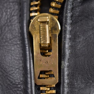 May include: Close-up of a gold-colored zipper pull with the word "TALON" embossed on it. The zipper pull is attached to a black leather jacket.
