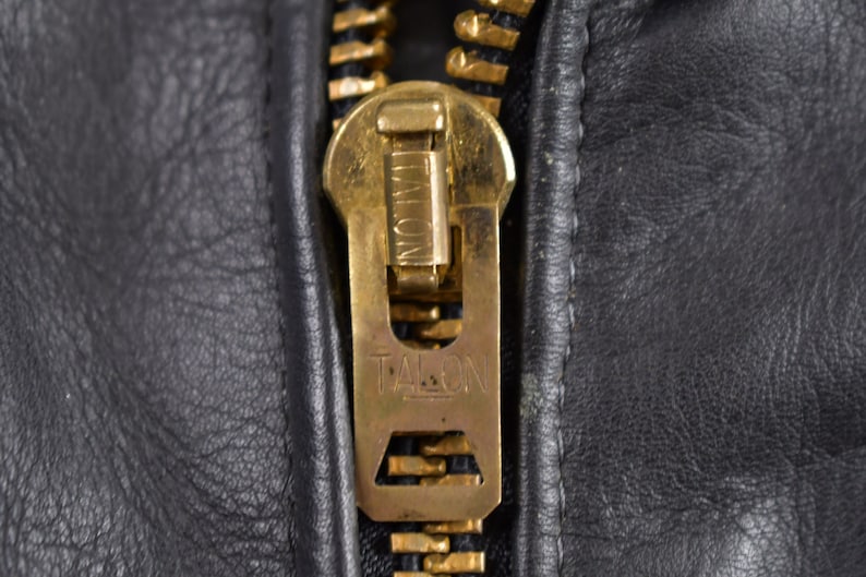 May include: Close-up of a gold-colored zipper pull with the word "TALON" embossed on it. The zipper pull is attached to a black leather jacket.