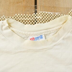 May include: Cream-colored, short-sleeved t-shirt with a crew neck. The Hanes brand tag is visible on the collar. The shirt is made of a soft, likely cotton material. The background is a wooden surface.