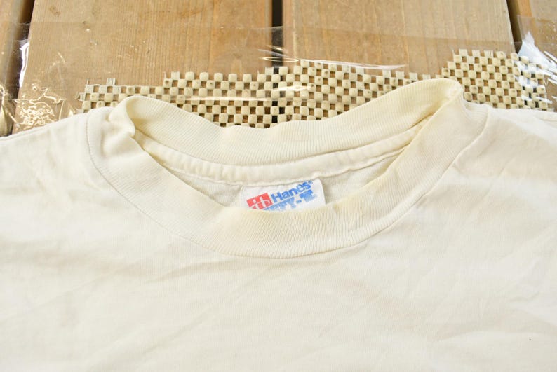 May include: Cream-colored, short-sleeved t-shirt with a crew neck. The Hanes brand tag is visible on the collar. The shirt is made of a soft, likely cotton material. The background is a wooden surface.