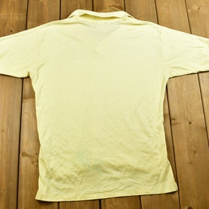 K&ouml;nnte beinhalten: Ein hellgelbes, kurz&auml;rmeliges Poloshirt mit Kragen. Das Shirt liegt flach auf einer Holzoberfl&auml;che.