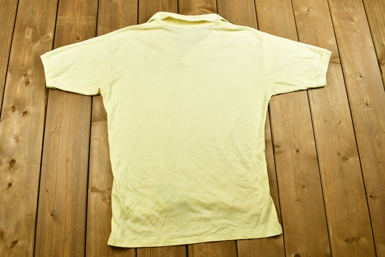 K&ouml;nnte beinhalten: Ein hellgelbes, kurz&auml;rmeliges Poloshirt mit Kragen. Das Shirt liegt flach auf einer Holzoberfl&auml;che.