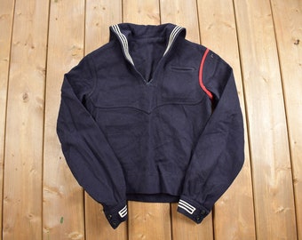 May include: A dark blue long-sleeved sweater with a white sailor collar and red trim on the right shoulder. The sweater has a V-neck and two buttons on each cuff.