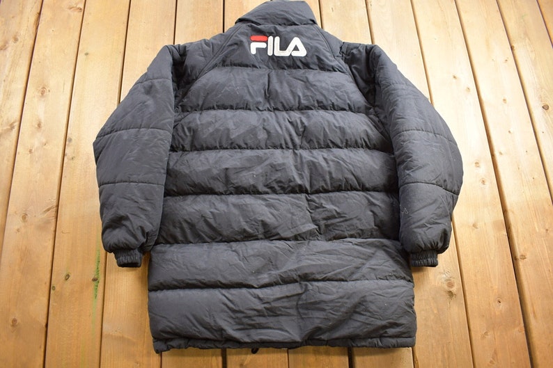 May include: Black puffer jacket with a FILA logo on the back. The jacket has a zip fastening and a stand-up collar.