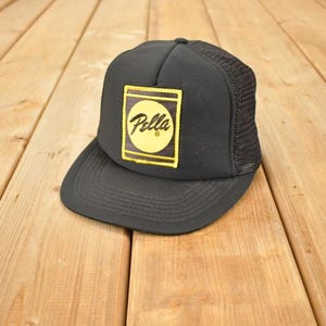 May include: Black trucker hat with a yellow and black logo that reads "Pella". The hat has a mesh back and a curved brim. The hat is sitting on a wooden surface.