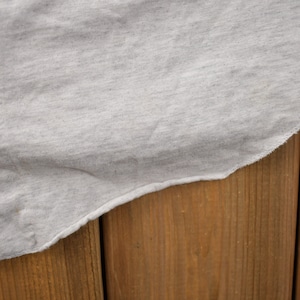 May include: A close-up of a light gray cotton shirt with a single button on the left side. The shirt is draped over a wooden surface.