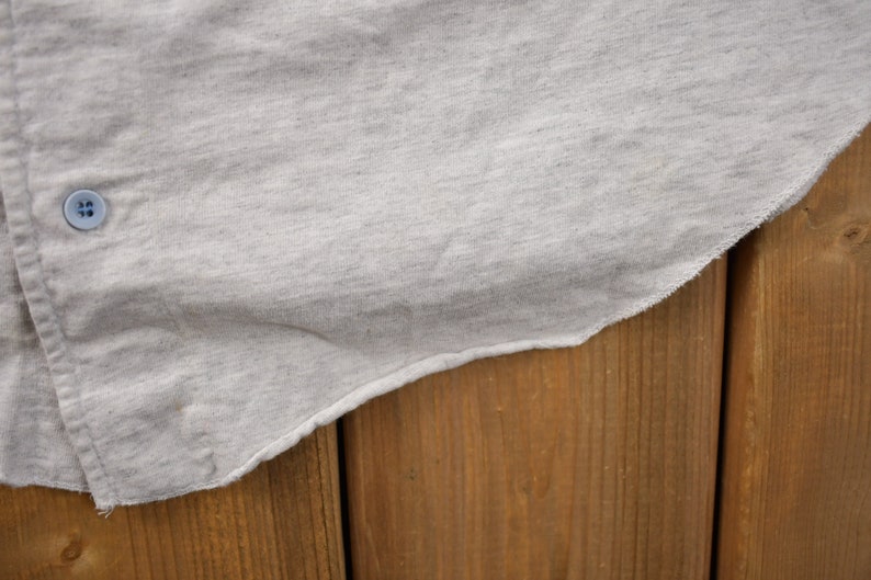 May include: A close-up of a light gray cotton shirt with a single button on the left side. The shirt is draped over a wooden surface.