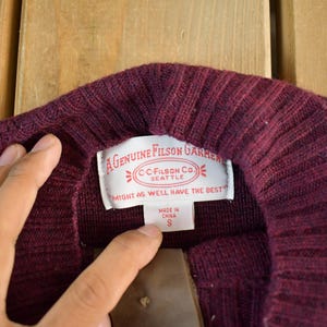 Puede incluir: Primer plano de un cuello de su&eacute;ter burdeos con textura acanalada. Una etiqueta blanca muestra el texto "A Genuine Filson Garment" y "Might as well have the best". La etiqueta tambi&eacute;n indica la talla S.